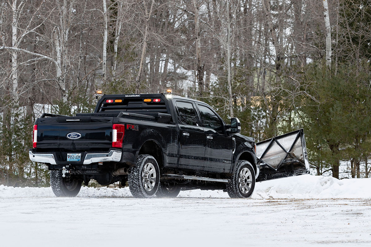 Black pickup driving over snowy ground with plow and Magnum headache rack installed